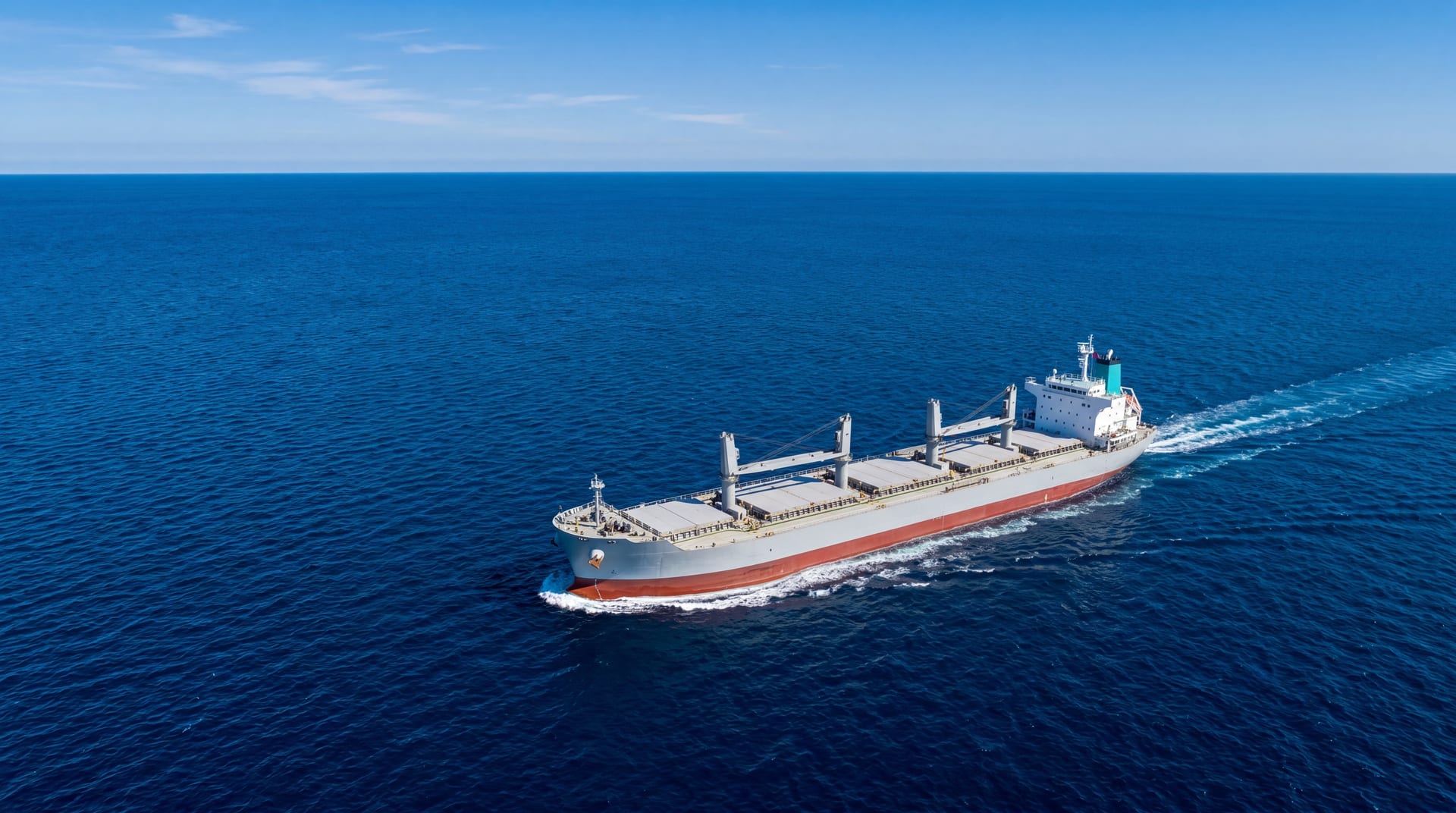 Aerial of a single modern bulk carrier cutting through clean deep blue ocean at midday, no exhaust trail, pristine waters