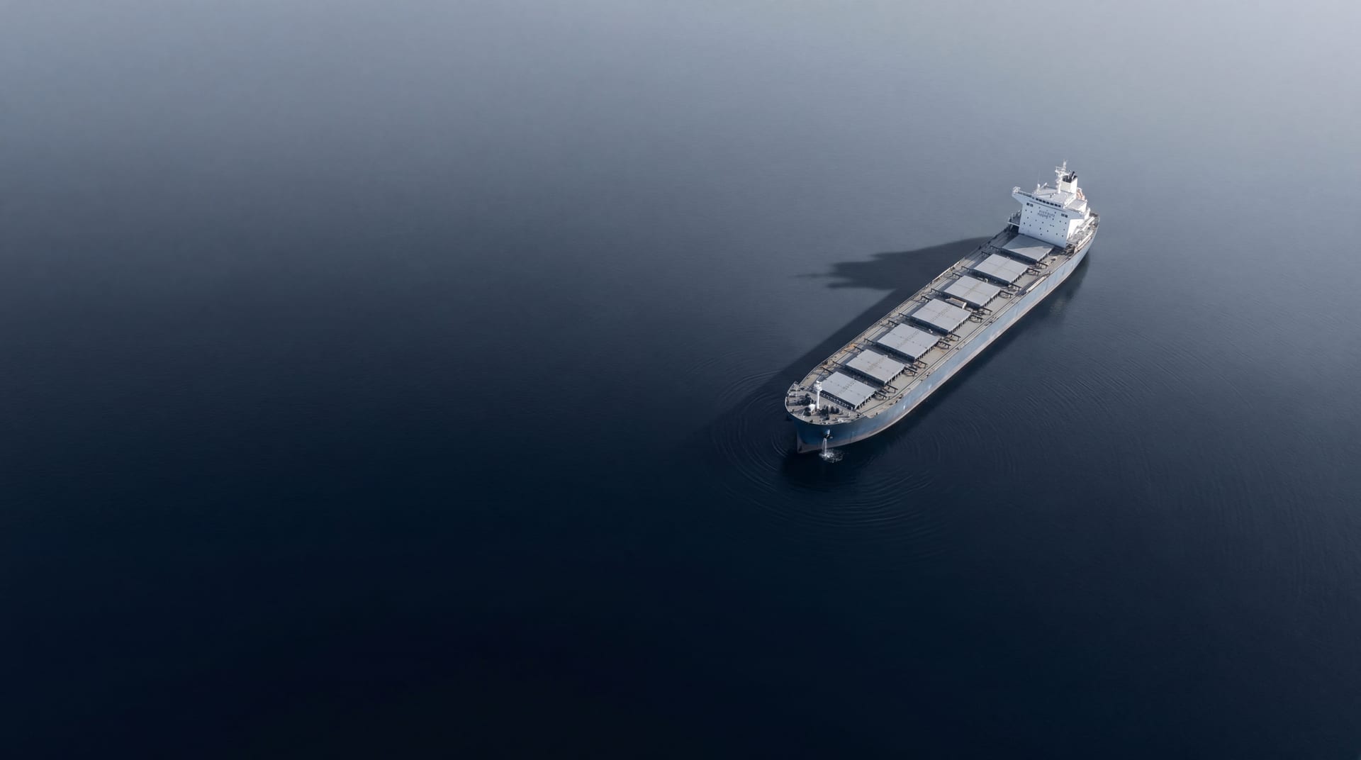 Single bulk carrier anchored alone in deep navy water, wide aerial with vast negative space